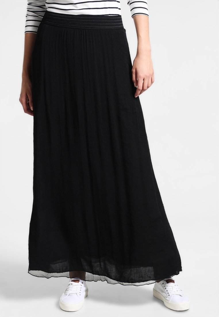 Femme portant une longue jupe plissée noire, des baskets blanches et un haut à manches longues blanc et noir à rayures, debout devant un fond blanc.