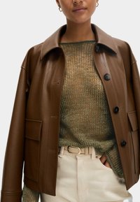 Veste en cuir marron avec un col, des boutons à l'avant et une poche à rabat, superposée à un pull en maille vert et un pantalon crème.