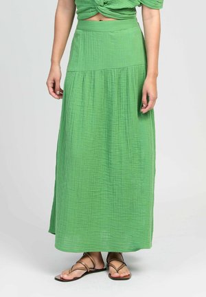 Jupe longue verte texturée portée avec des sandales plates à brides marron, visible de la taille aux pieds sur un fond uni.