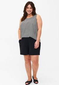 Top senza maniche a motivi in bianco e nero con orlo curvato, abbinato a pantaloni corti neri. Il modello posa con le mani in tasca, indossando sandali neri.