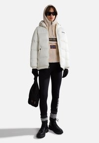 Giacca puffer bianca indossata sopra un felpa beige di pile con la bandiera norvegese, leggings neri, calzini con dettagli grigi e stivali militari neri.