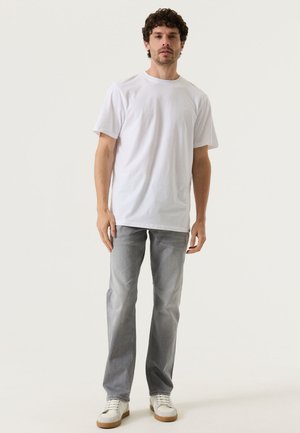 Ein Mann steht und trägt ein schlichtes weißes T-Shirt, hellgraue Jeans und weiße Sneakers vor einem schlichten hellen Hintergrund.