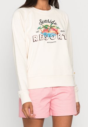 Kvinde iført cremefarvet sweatshirt med farverigt motiv af kystresort og rosa shorts, vist fra skuldre til midt på låret.
