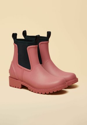 Pereche de ghete Chelsea din cauciuc roz mat, cu panouri elastice laterale negre și barete pentru tras, pe un fundal bej.