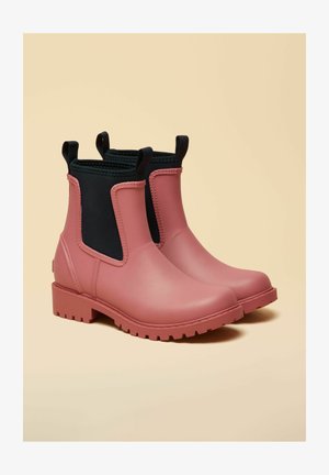 Par af matte lyserøde gummichelsea-støvler med sorte elastiske sidepaneler og trækflapper, sat mod en beige baggrund.
