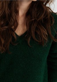 Grüner, strukturierter Pullover mit V-Ausschnitt. Das Material wirkt weich und gestrickt, mit einem groben Maschenbild. Hintergrund teilweise durch Haare verdeckt.