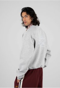 Felpa grigia con polsini a coste, con un logo bianco ricamato sulla schiena. Abbinata a pantaloni bordeaux con strisce laterali.