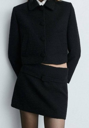 Vrouw draagt een zwarte, gestructureerde cropped jas met grote knopen en een bijpassende minirok met hoge taille en brede tailleband.