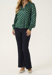 Blusa verde con motivi ovali dorati, scollo a v e maniche lunghe, abbinata a jeans svasati scuri e scarpe con tacco beige.