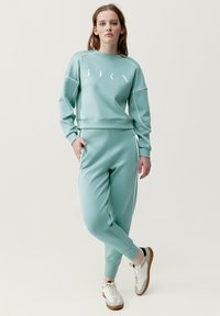 Felpa sportiva verde menta chiaro con dettagli bianchi e logo, abbinata a pantaloni della tuta coordinati con design a gamba affusolata.