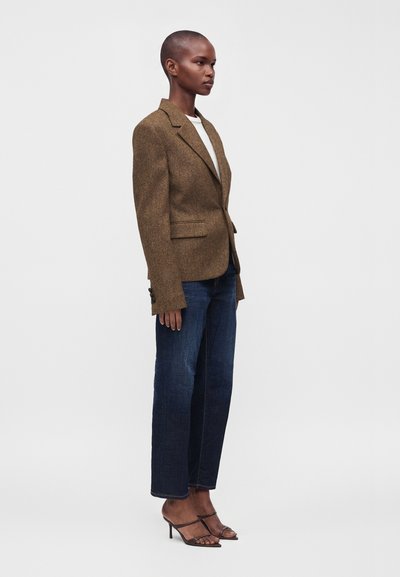 Femme debout de profil portant un blazer marron, un haut blanc, un jean bleu foncé et des sandales marron à talons sur un fond uni.