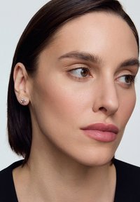 Srebrne kolczyki w kształcie kwiatu z małymi akcentami kryształowymi. Zbliżenie na ucho modelki, podkreślające wzór i blask kolczyków.