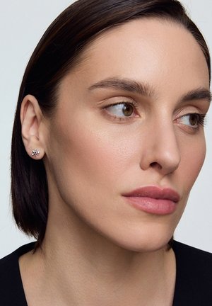 Srebrne kolczyki w kształcie kwiatu z małymi akcentami kryształowymi. Zbliżenie na ucho modelki, podkreślające wzór i blask kolczyków.