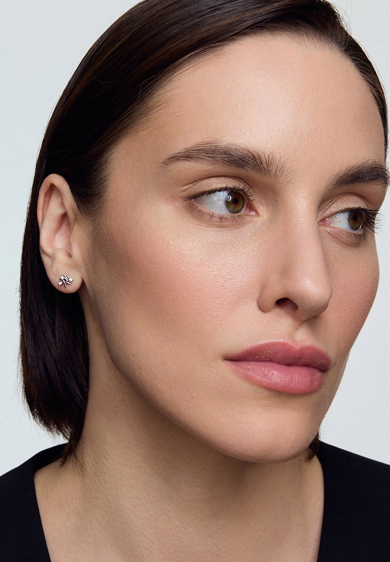 Srebrne kolczyki w kształcie kwiatu z małymi akcentami kryształowymi. Zbliżenie na ucho modelki, podkreślające wzór i blask kolczyków.