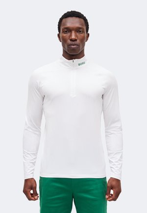 Bărbat purtând o cămașă albă cu mâneci lungi cu fermoar și „BORG” pe guler și pantaloni verzi, stând în fața unui fundal simplu.