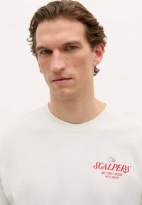 Sweatshirt blanc avec un col rond, des poignets côtelés et un logo rouge brodé sur la poitrine gauche, avec une texture lisse et douce.