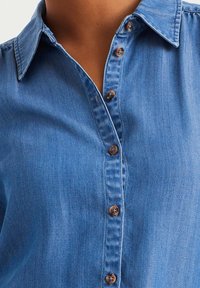 Blue denim shirt with a classic collar, seven button front closure, and textured fabric. Buttons are brown with a slight sheen.