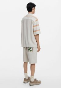 Korte mouwen overhemd met horizontale strepen in beige, oranje en blauw; gecombineerd met lichtbeige cargoshorts met palmbomen borduursel.