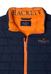 Marinblå puffjacka med orange detaljer, med horisontell quiltning, dragkedja fram och "Hackett"-logotyp på kragen och bröstet. Storlek 7-8 år.