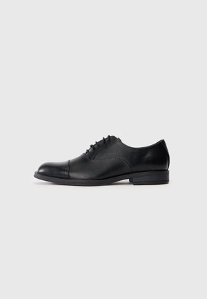 Black leather men's oxford shoe with closed lacing, cap toe, and low stacked heel, shown in side profile on light gray background.