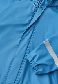 Lysblå, vandtæt jakke med en glat tekstur. Har lynlåslukning og elastiske manchetter. Bemærkelsesværdi sølvaccent på ærmet.