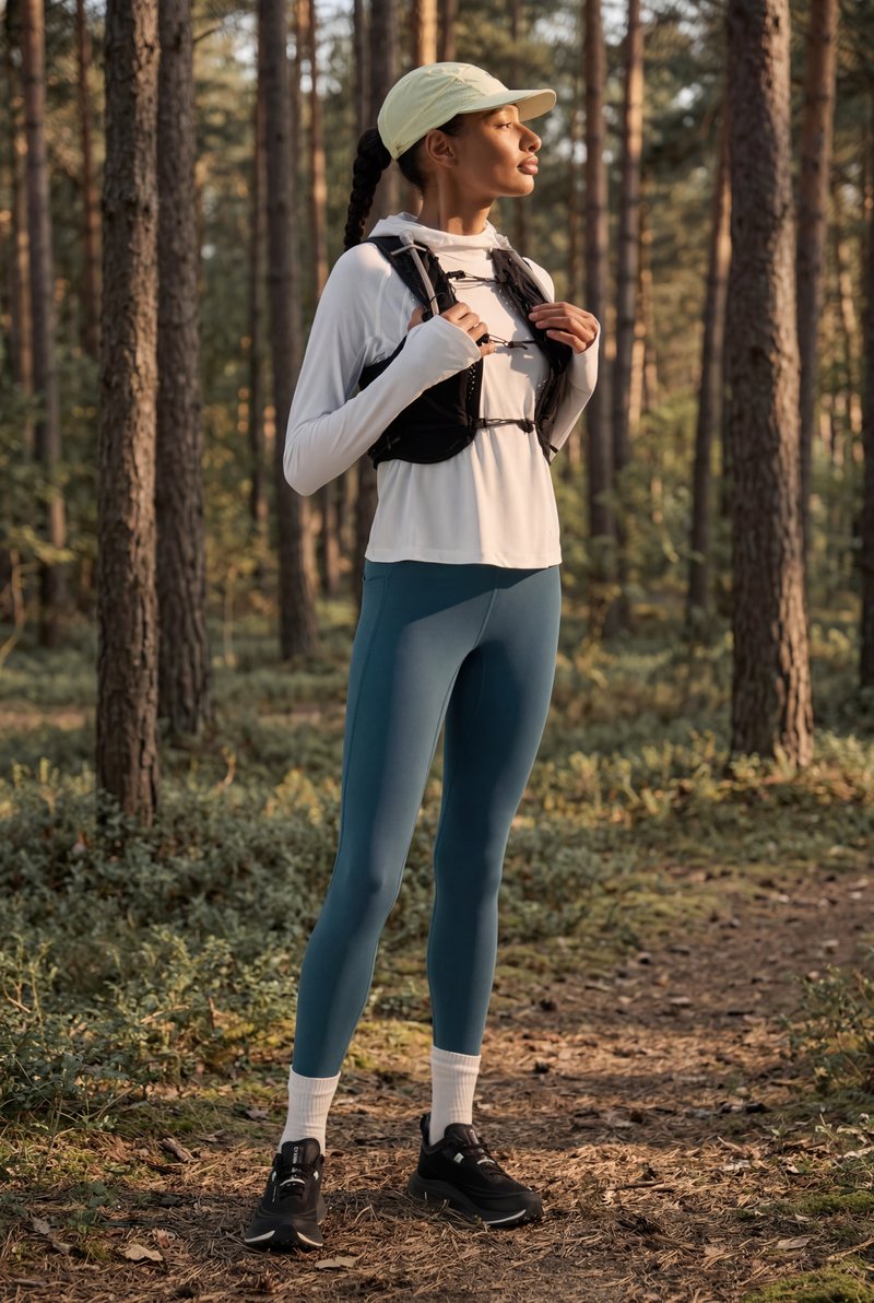 Žena u odjeći za trčanje podešava prsluk za hidrataciju, stoji na šumskoj stazi okružena visokim drvećem u mekanom sunčevom svjetlu.