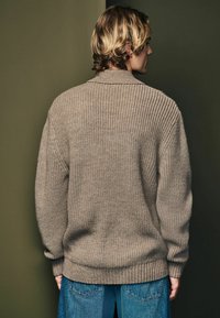 Pull en tricot beige avec un col et des poignets côtelés, présentant un motif texturé. Porté avec un jean en denim bleu. Fond en vert atténué.