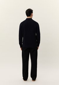 Άνδρας που στέκεται με την πλάτη, φοράει μαύρο ριμπ πλεκτό πουλόβερ με μακριά μανίκια και φαρδιά μαύρη παντελόνια, μπροστά σε ένα απλό ανοιχτό φόντο.