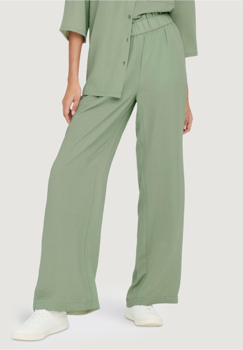Personne portant un pantalon ample vert clair avec une ceinture élastique et des baskets blanches, debout devant un fond uni.