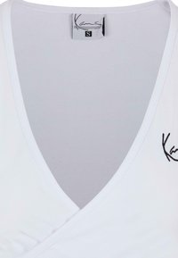 Biele zapínacie tričko s hlbokým výstrihom do V, so čiernym vyšívaným logom na ľavej strane. Vyrobené z hladkej, elastickej látky s elegantným povrchom.
