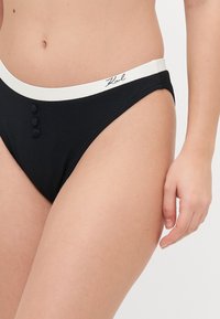 Black bikini bottom featuring a textured fabric, high-cut leg openings, a white waistband with buttons, and "Karl" logo on the front.