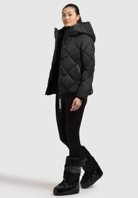 Giacca piumino nera con design a rombi e cappuccio grande, abbinata a leggings neri e stivali invernali lucidi. Presenta zip e marchio.