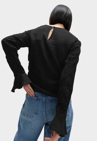 Svart topp med texturerat tyg, långa ärmar och utsvängda manschetter; har en nyckelhålsdetalj bak. Matchas med blå denimjeans.