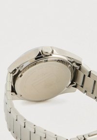 Κοντινό πλάνο ενός ασημένιου κομμένο από ανοξείδωτο ατσάλι λουριού ρολογιού και πίσω πλευράς, που δείχνει την κορώνα και τις χαραγμένες επιγραφές της μάρκας.