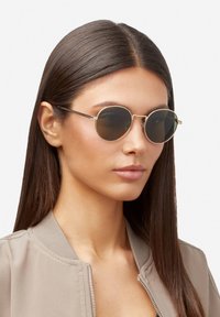 Gafas de sol redondas con lentes oscuros y delgadas monturas de metal dorado. El modelo tiene el cabello largo y liso de color castaño y lleva puesta una chaqueta bomber beige.