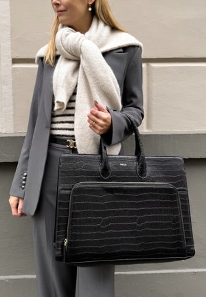 Black leather bag with a crocodile texture, featuring a zippered front pocket and dual handles, held by a person in a gray suit and scarf.