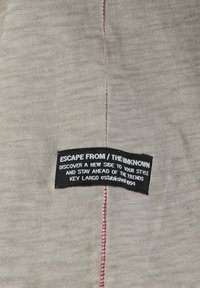 Tejido gris claro con una superficie ligeramente texturizada, que presenta una etiqueta negra prominente con texto blanco y costura roja a lo largo de la costura.