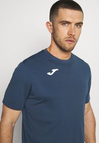 Sötétkék, rövid ujjú sportpóló sima anyagból, bal oldalon fehér logóval, kerek nyakkivágással.