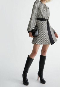 Donna che indossa un abito a fantasia bianco e nero con maniche lunghe, una cintura nera, stivali neri fino al ginocchio e tiene in mano una piccola borsa nera.