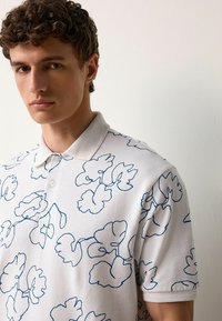 Cămașă polo albă, realizată dintr-un material texturat, cu contururi florale albastre ca model. Include o guler și închidere cu nasturi.