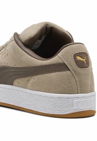 Suede sneakers in beige with dark brown accents, featuring a textured white midsole and a gum rubber outsole. Notable Puma logo on heel.