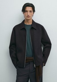 Donker, gestructureerd jasje met een kraag en rits, gedragen over een groene blouse en een bruine wikkel, in combinatie met donkere broeken. Minimalistisch ontwerp.