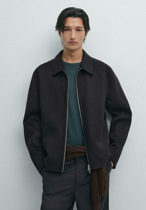 Dark, textured jacket with a collar and zipper, worn over a green shirt and brown wrap, paired with dark trousers. Minimalist design.