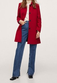 Femme portant un manteau rouge à boutons sur un col roulé blanc, un jean bleu taille haute avec fentes à l'avant et des chaussures noires à bout carré.