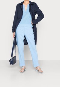 Cappotto impermeabile blu navy sopra un maglione azzurro chiaro, abbinato a pantaloni azzurri chiari e sandali coordinati. Piccola borsa a tracolla blu scuro.