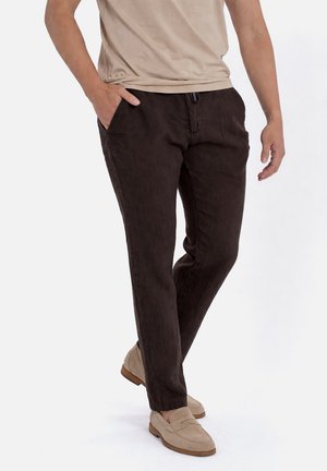 Man draagt een beige shirt met korte mouwen, donkerbruine broek en lichtbeige loafers, staand met één hand in de zak tegen een witte achtergrond.