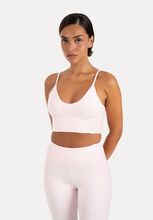 Kvinde med kort mørkt hår iført en lyserød sports-bh og matchende højtaljede leggings, der står foran en ensfarvet baggrund.