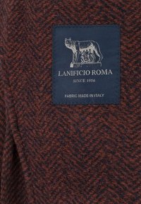 Strukturalny materiał z wzorem w rybi ogon w brązowym i granatowym kolorze, z niebieską metką z grafiką wilka i napisem "LANIFICIO ROMA".