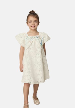 Jong meisje dat een knie-lange witte kanten jurk draagt met wijde mouwen en een grote bloemige strik op de borst, glimlachend en staand.