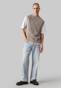 Maglione smanicato beige intrecciato sopra una T-shirt bianca, abbinato a jeans a gamba dritta blu chiaro e scarpe nere. Design semplice e texture.